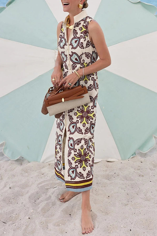 Woman in a patterned dress standing on a sandy beach with a colorful umbrella in the background.